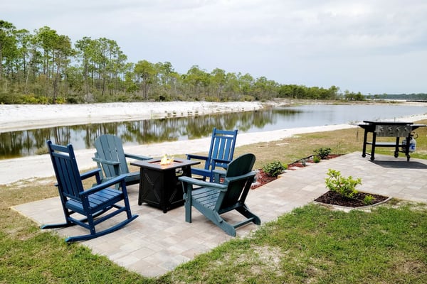 Waterfront patio with blue Adirondack chairs, fire pit table, and grill overlooking calm bay and pine forest.