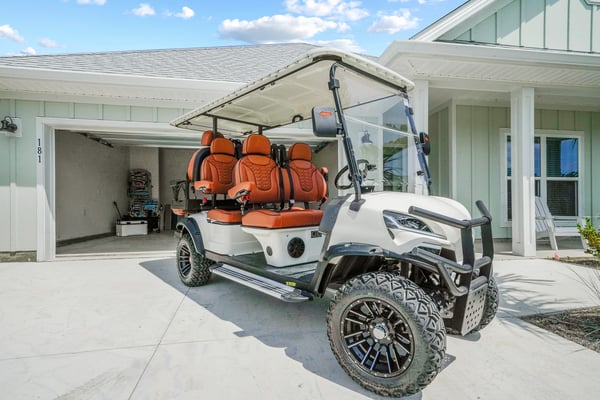 White and teal golf cart with orange seats parked in driveway of modern coastal home with open garage.